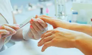 Dłoń klienta z pomalowanymi paznokciami jest piłowana przez kosmetyczkę w rękawiczkach.