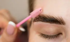 Zbliżenie na oko, gdzie różowa szczoteczka do brwi stylizuje ciemne brwi.