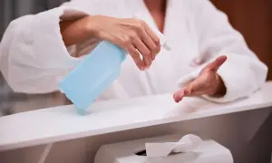 Dłoń w białym szlafroku trzyma niebieski spray dezynfekujący nad białą powierzchnią.