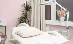 Biały fotel kosmetyczny z poduszką i ręcznikiem, lampa, roślina i dekoracje na jasnoróżowym tle.