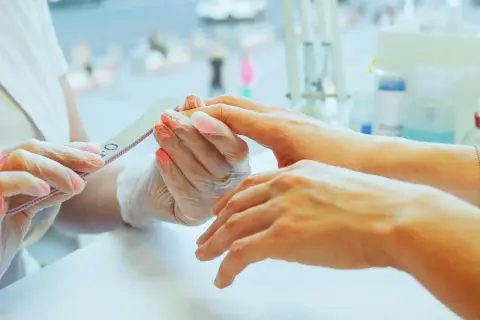 Dłoń klienta z pomalowanymi paznokciami jest piłowana przez kosmetyczkę w rękawiczkach.