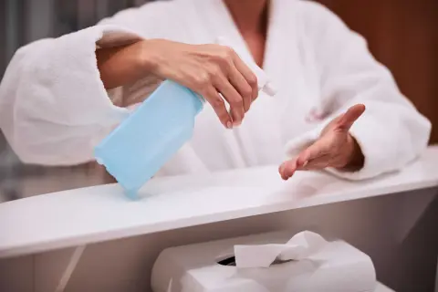 Dłoń w białym szlafroku trzyma niebieski spray dezynfekujący nad białą powierzchnią.