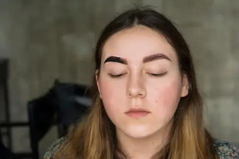 Dziewczyna z zamkniętymi oczami, na jednej brwi ciemna pasta, druga już pomalowana. Efekt pudrowe brwi w trakcie tworzenia.