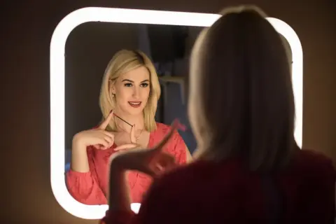 Kobieta z blond włosami w czerwonej bluzce patrzy w lustro, trzymając szczoteczkę do rzęs.