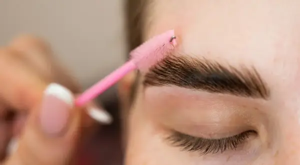 Zbliżenie na oko, gdzie różowa szczoteczka do brwi stylizuje ciemne brwi.