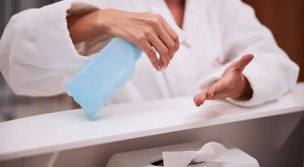 Dłoń w białym szlafroku trzyma niebieski spray dezynfekujący nad białą powierzchnią.