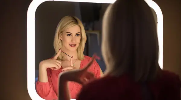 Kobieta z blond włosami w czerwonej bluzce patrzy w lustro, trzymając szczoteczkę do rzęs.