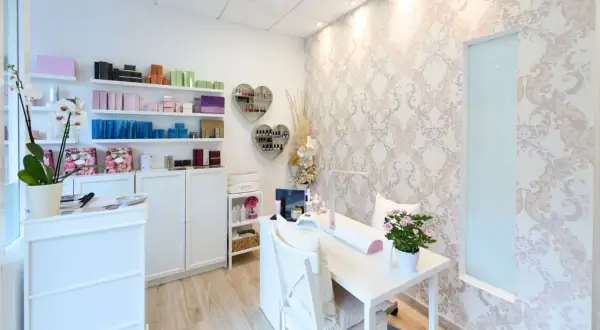 Jasny, schludny gabinet kosmetyczny z białymi meblami, półkami z produktami i stołem do manicure.