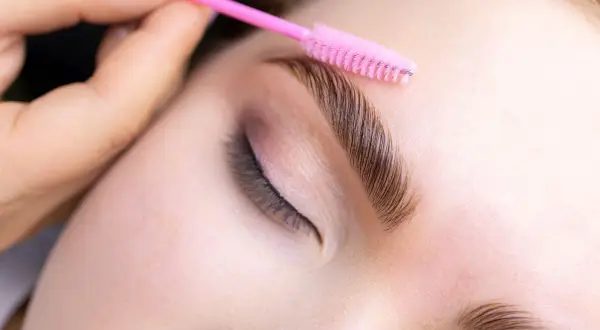 Stylist układa brwi po laminacji różową szczoteczką. Zastanawiasz się, jaki olejek po laminacji brwi będzie najlepszy?