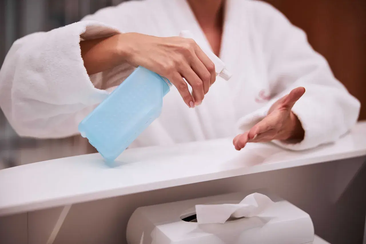 Dłoń w białym szlafroku trzyma niebieski spray dezynfekujący nad białą powierzchnią.