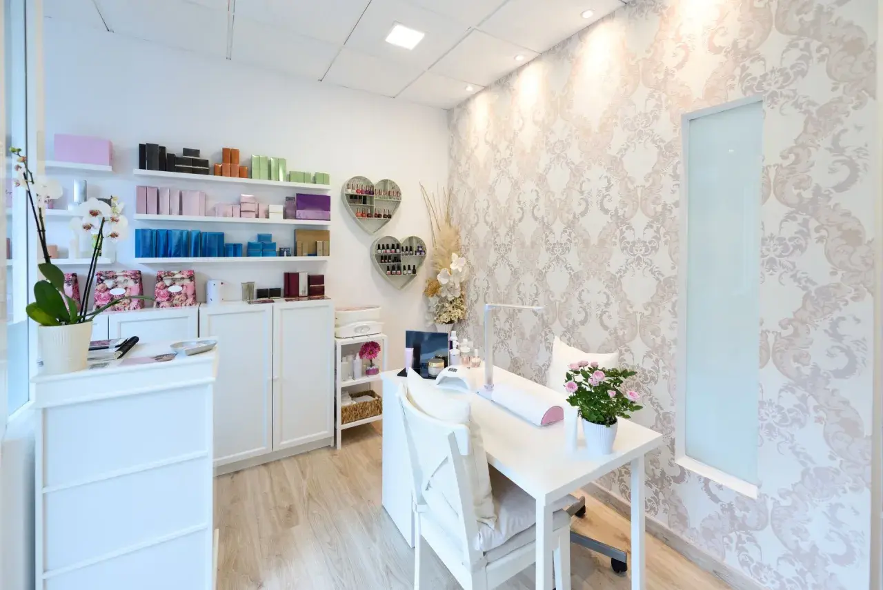 Jasny, schludny gabinet kosmetyczny z białymi meblami, półkami z produktami i stołem do manicure.