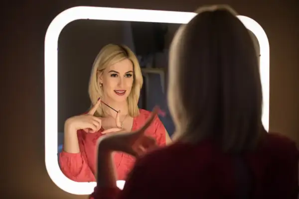 Kobieta z blond włosami w czerwonej bluzce patrzy w lustro, trzymając szczoteczkę do rzęs.