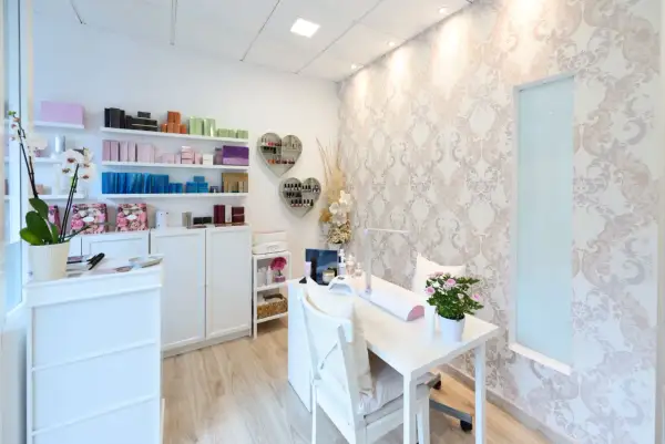 Jasny, schludny gabinet kosmetyczny z białymi meblami, półkami z produktami i stołem do manicure.